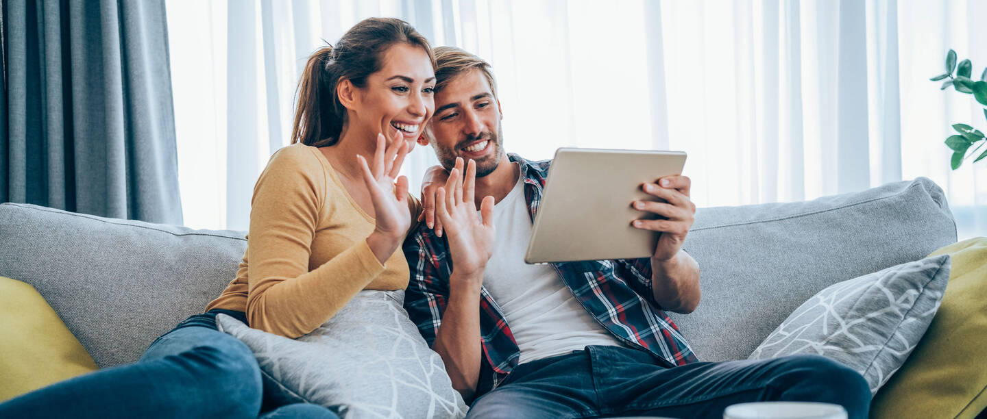 Ein junges Paar sitzt auf dem Sofa. Sie halten das Tablet in den Händen und winken es an.